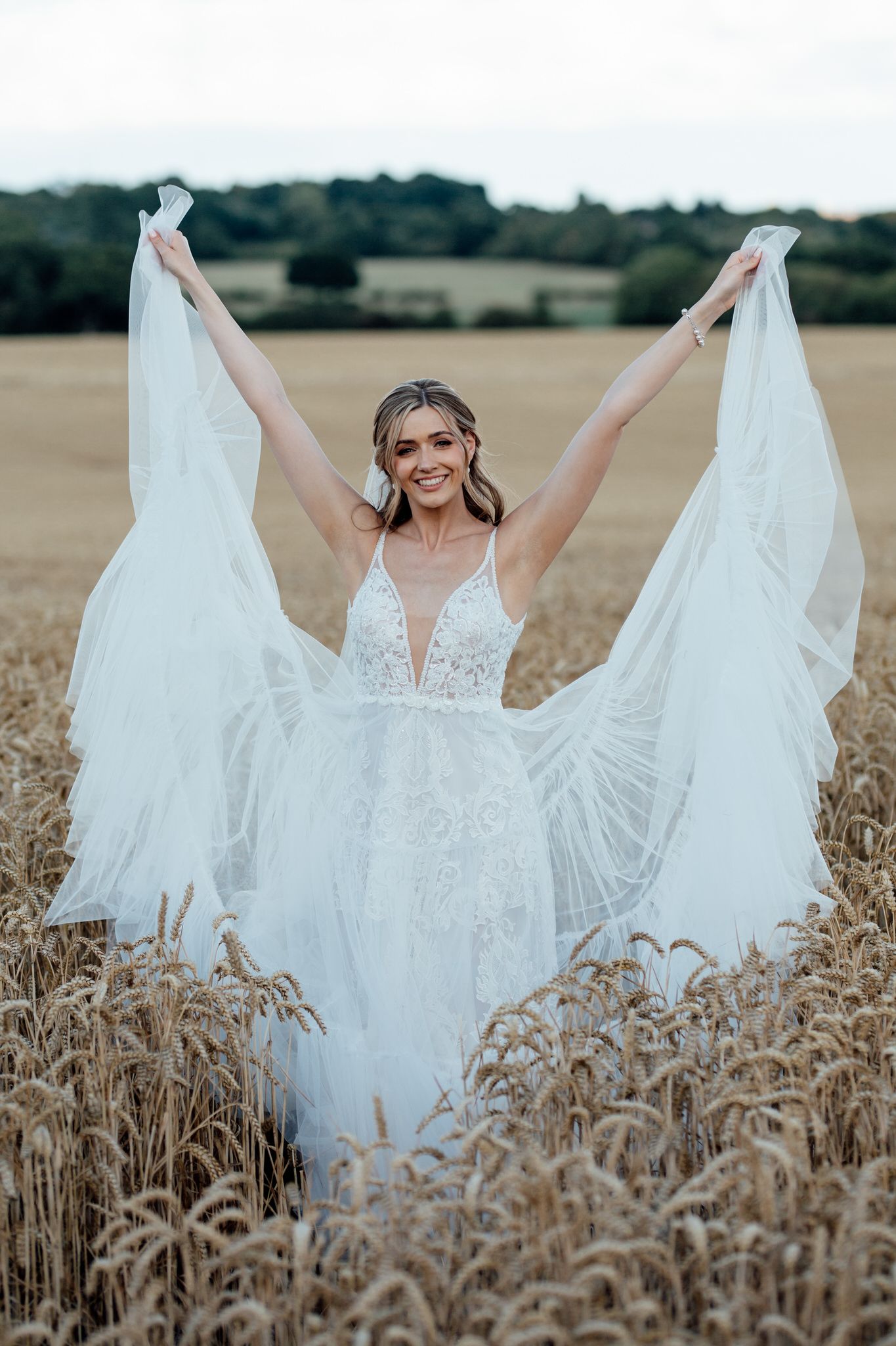 Bride in lace gown twirls veil dramatically while standing in golden wheat field.