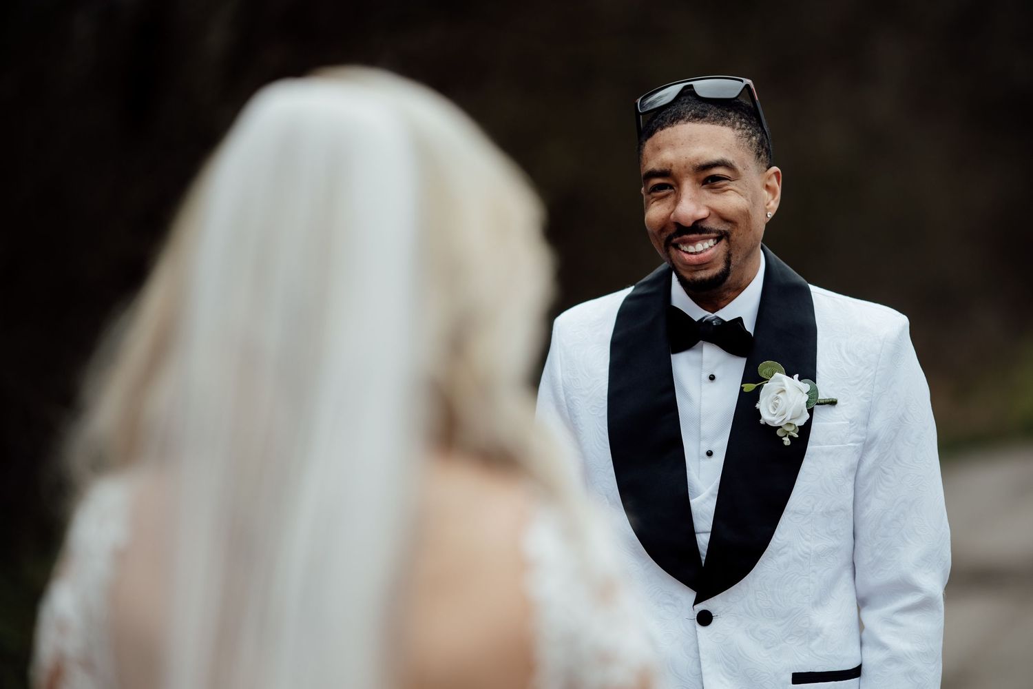 Groom in white tuxedo smiling at bride during outdoor wedding ceremony first look moment.