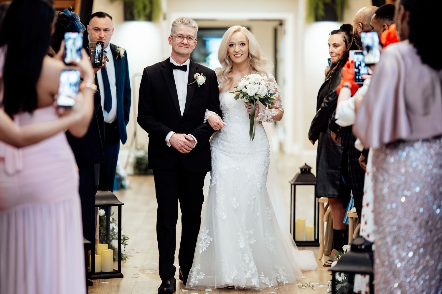 Bride in a white lace gown walks down the aisle with her father during an elegant indoor wedding ceremony.