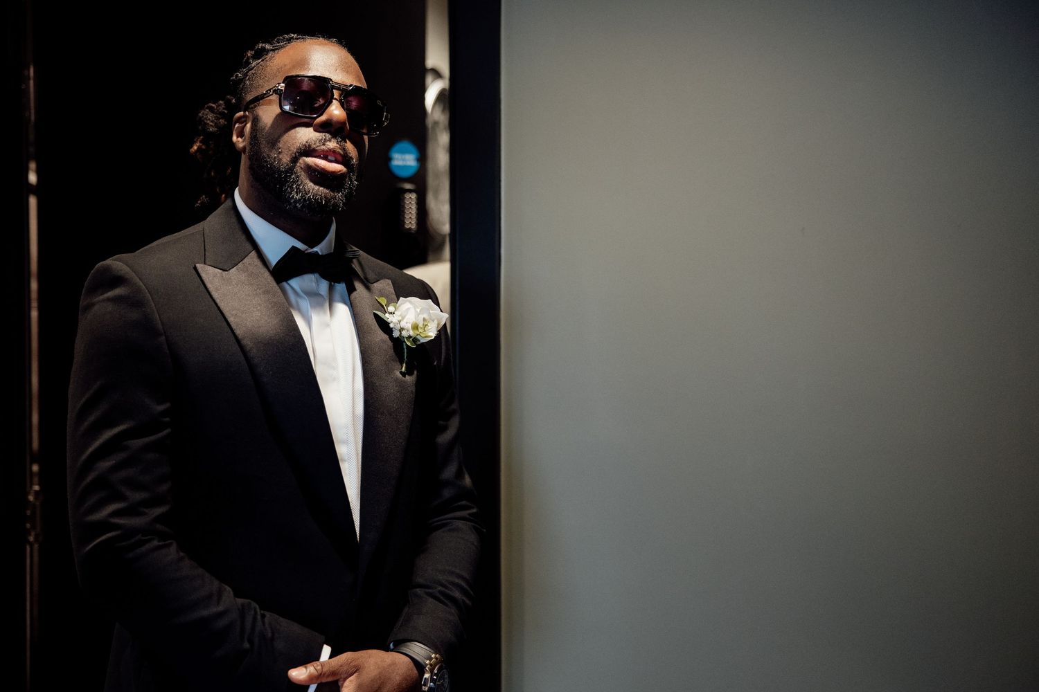 Stylish groom in black tuxedo with bow tie and sunglasses posing confidently before wedding ceremony.