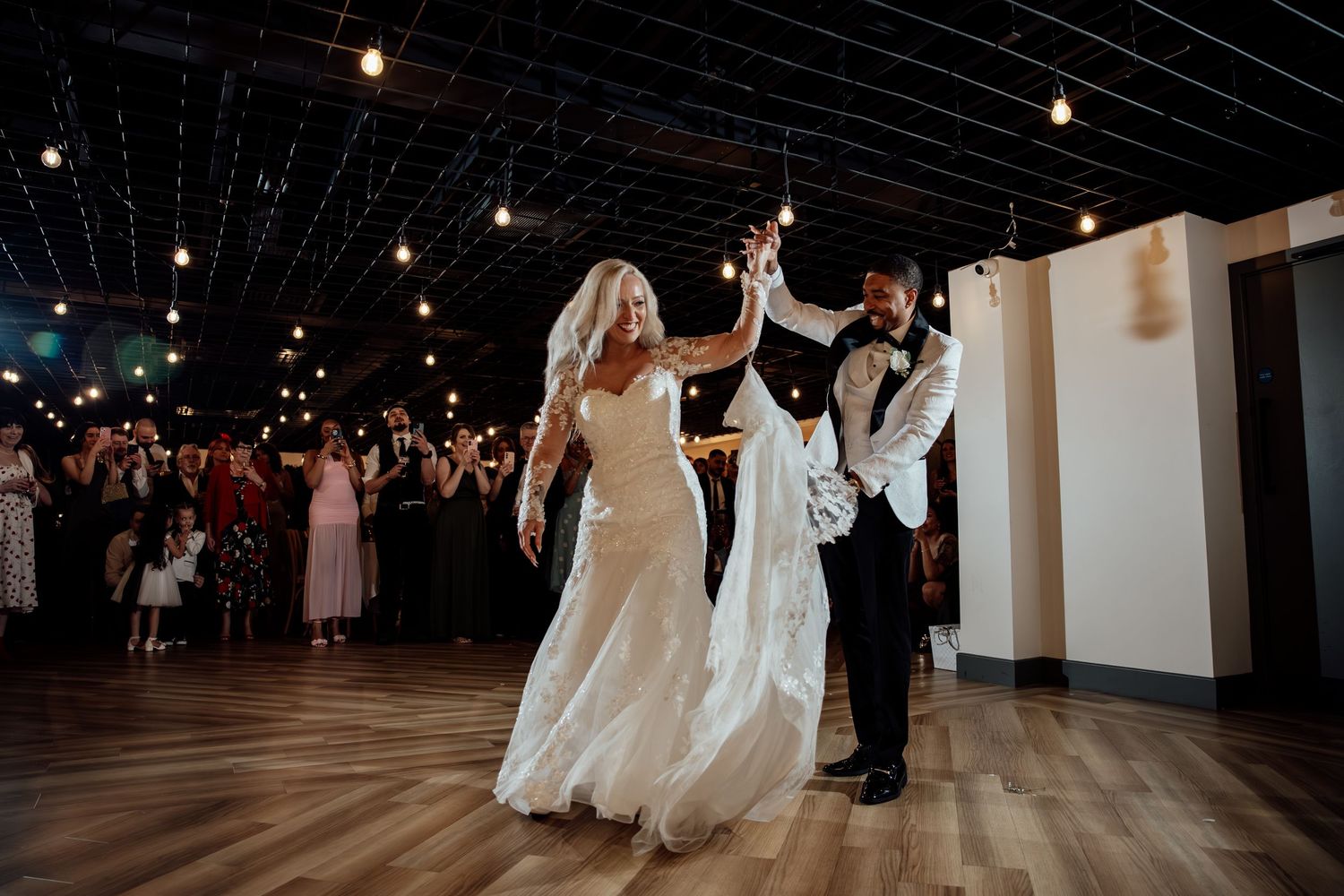 Bride and groom share their first dance under string lights surrounded by cheering wedding guests.