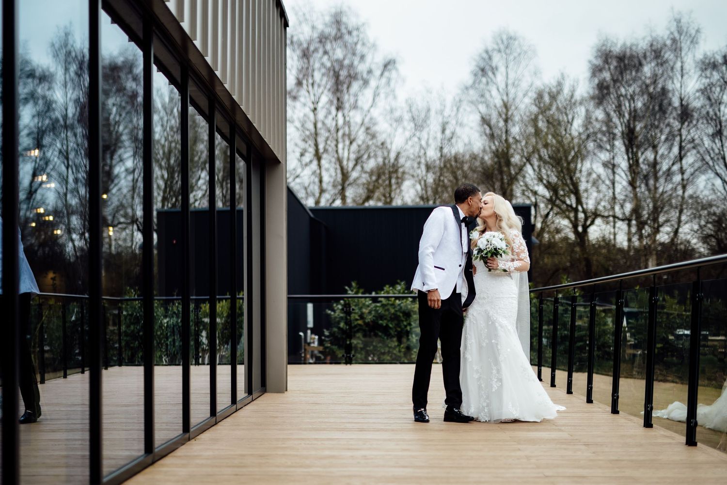 A bride and groom share a romantic kiss on a wooden deck surrounded by bare winter trees.