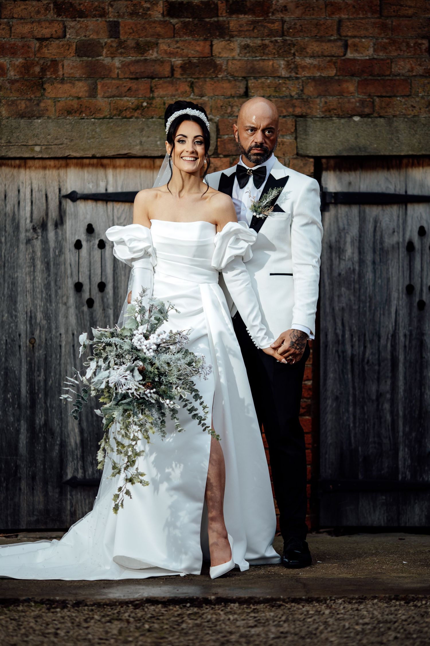 Bride in off-shoulder gown and groom in white tuxedo embrace near weathered wooden wall.