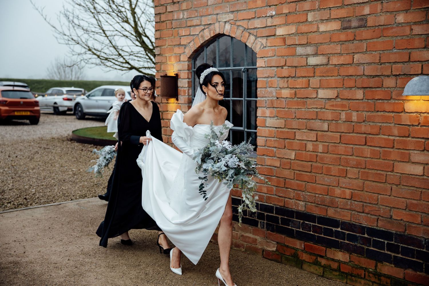 Wedding party members assist with dress outside brick building entrance.