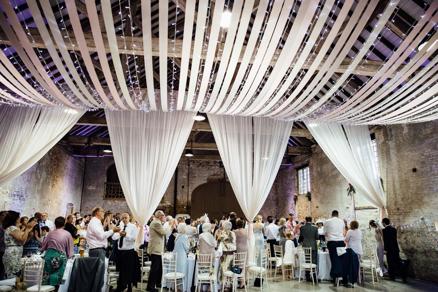 A wedding reception venue with white draping and string lights features guests seated at long tables.