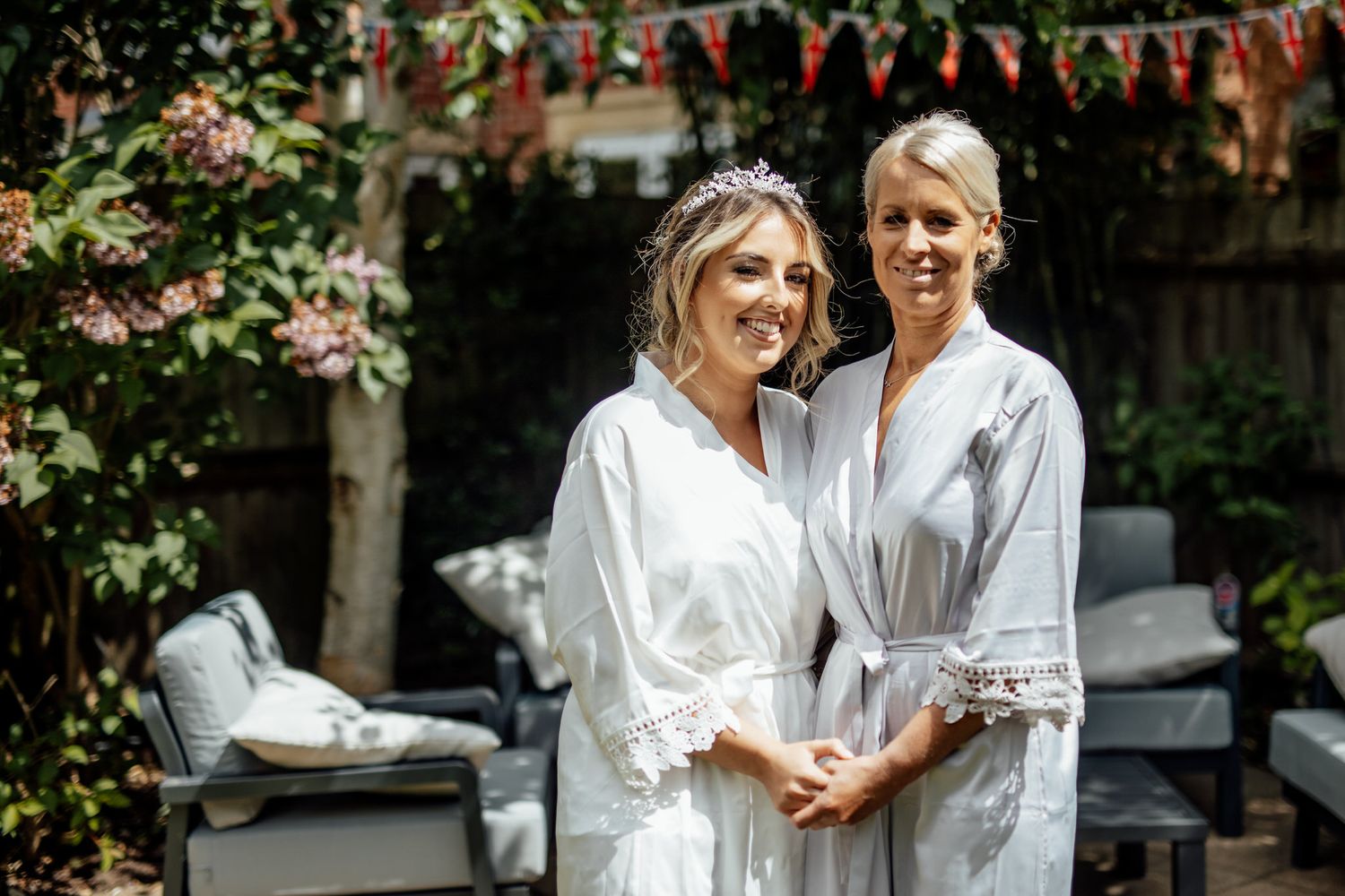 Two women in white robes posing in a garden setting with bunting decorations.