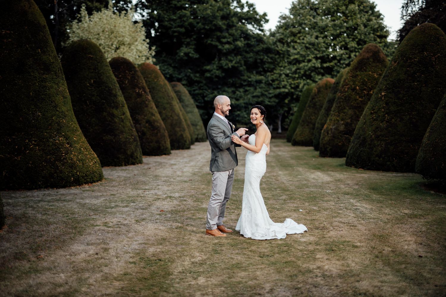 Wedding couple posing between rows of perfectly manicured topiary trees in formal garden.