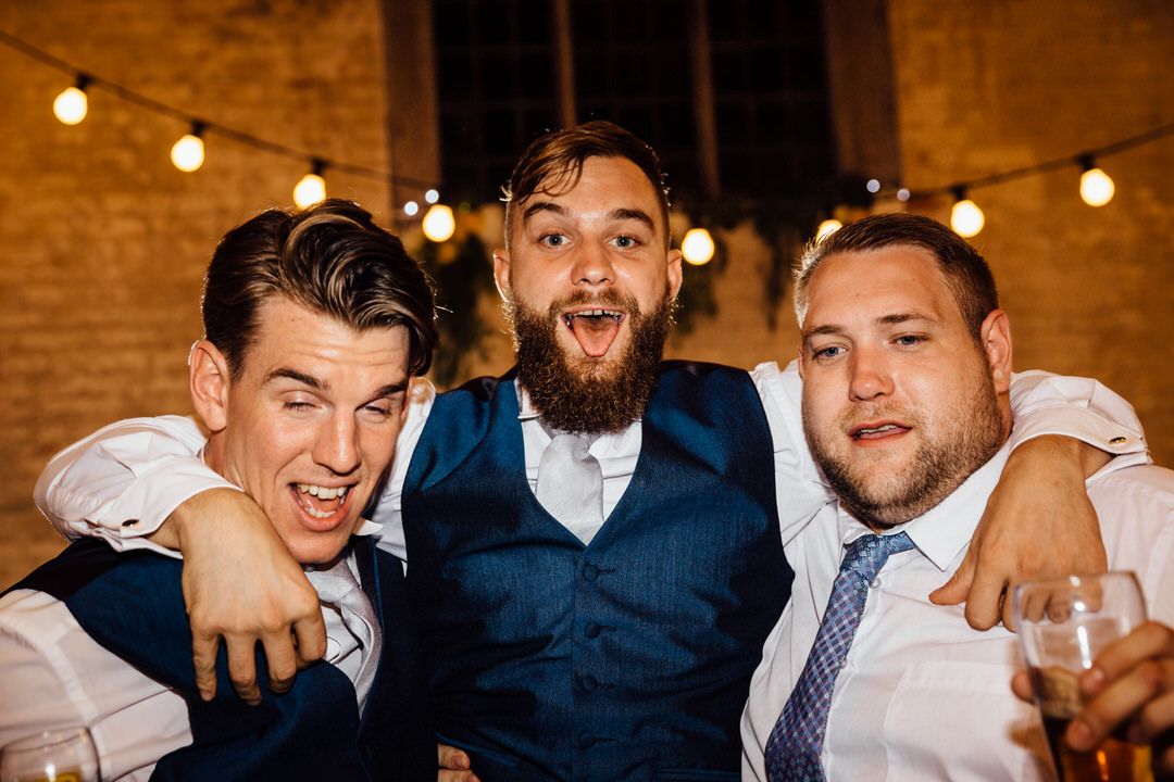 Groomsmen in navy suits and ties celebrate at rustic brick venue with string lights.