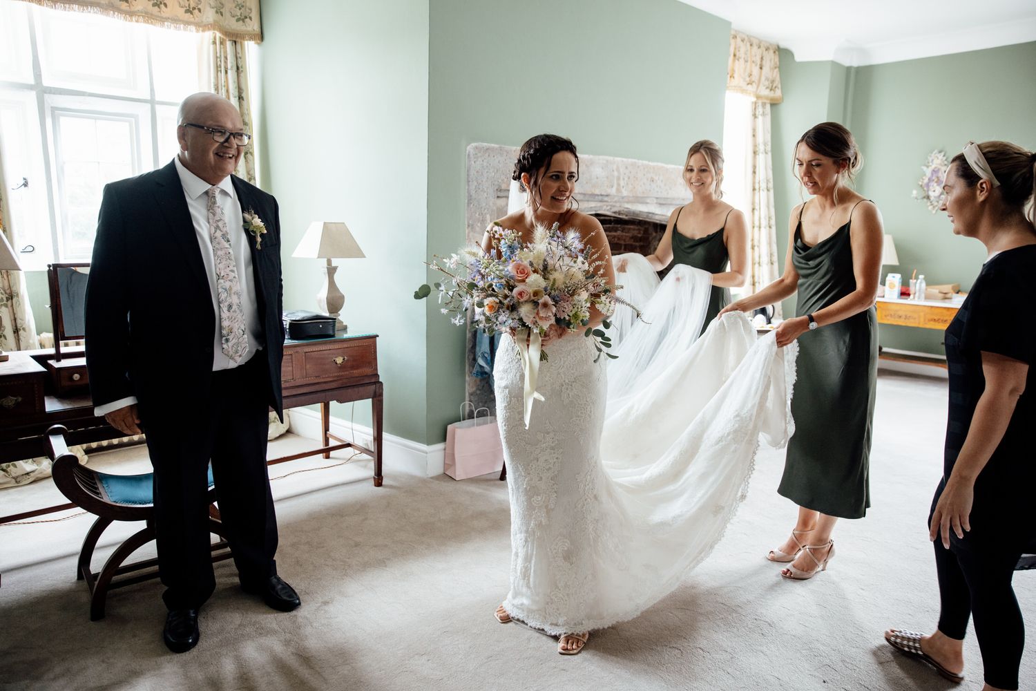 Wedding preparation scene in a mint green room with dress and attendants helping.