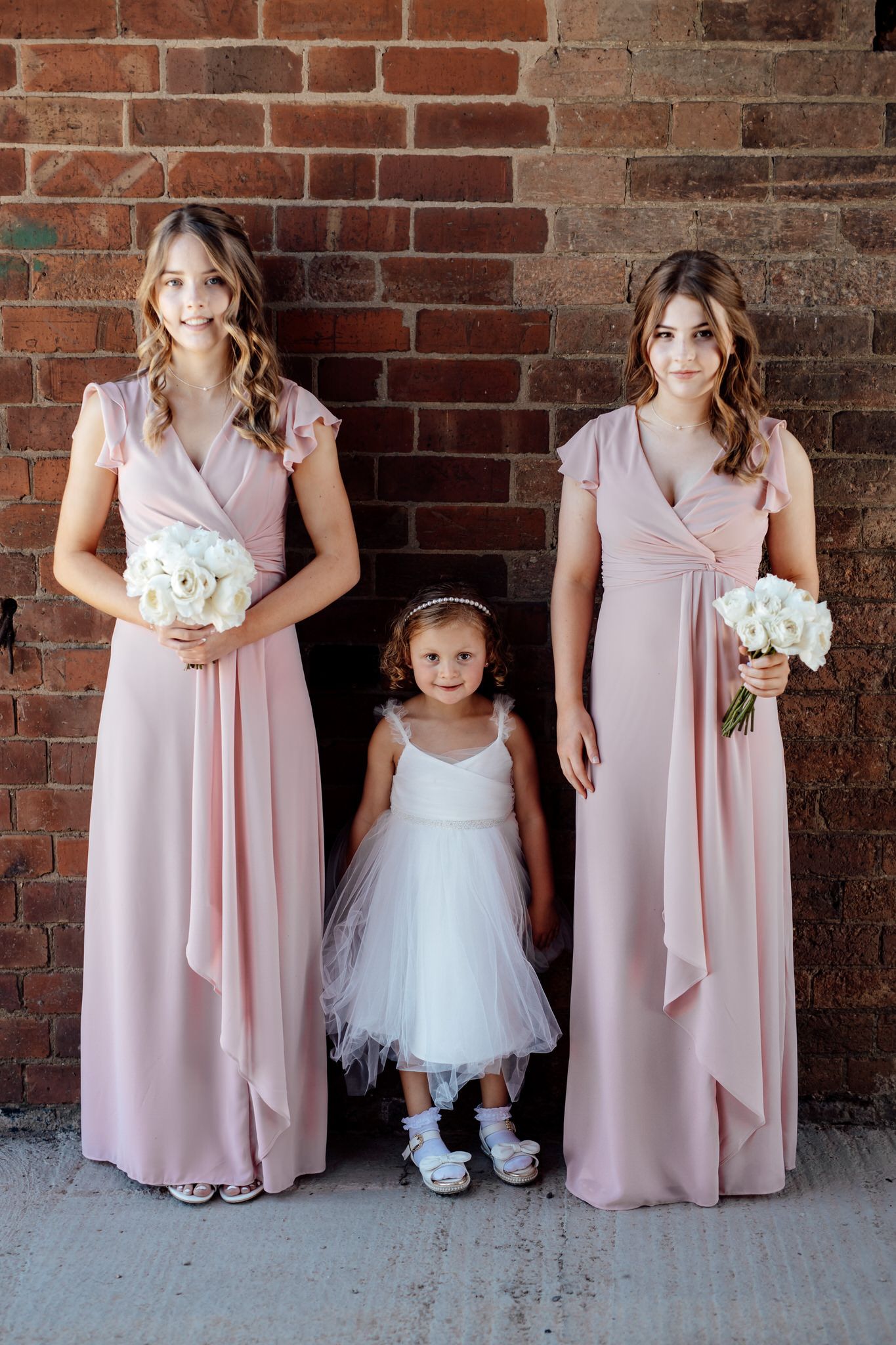 Bridesmaids in blush pink gowns and a flower girl in white tulle pose against a brick wall.
