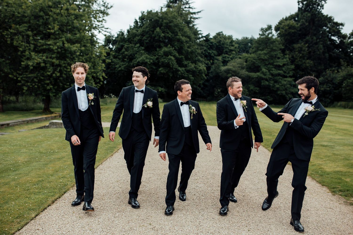 Groomsmen in black tuxedos walk together on a garden path while kicking their legs in unison.