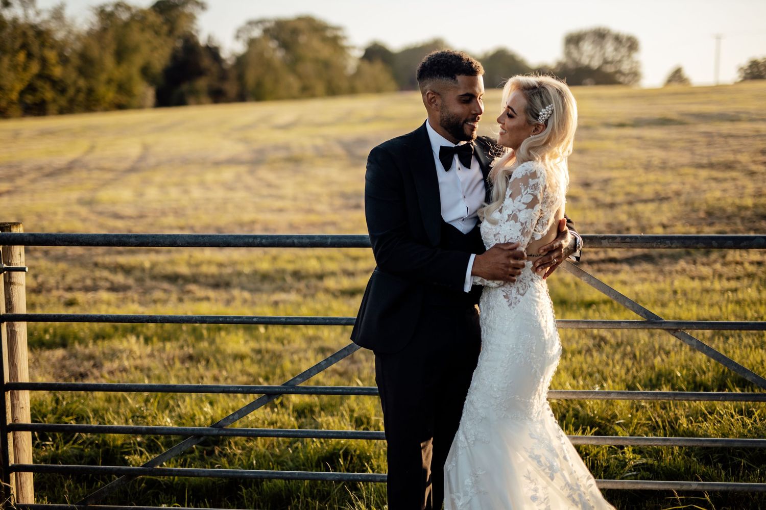 Newlyweds share a romantic moment along a rustic wooden fence at sunset.