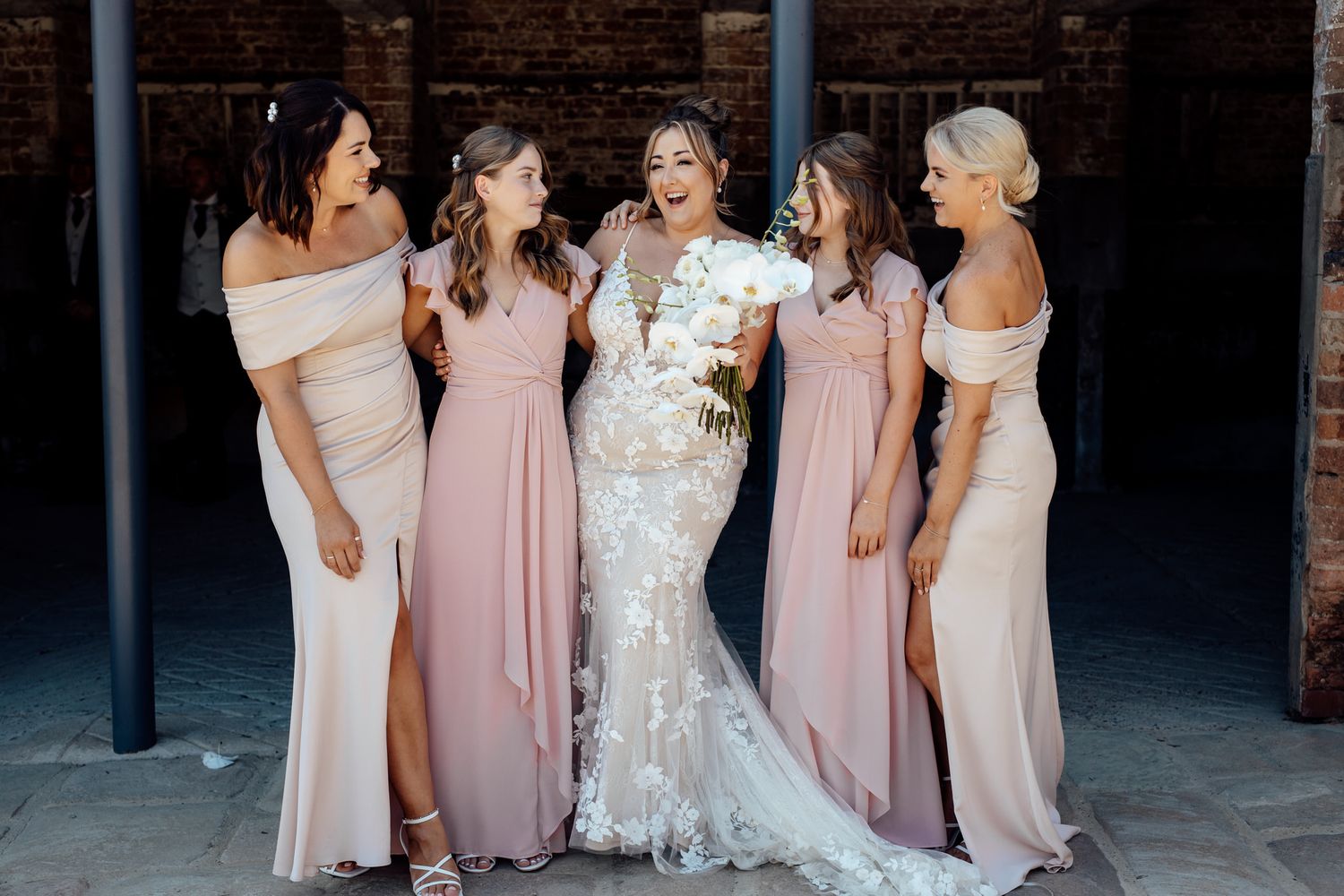 Wedding party in blush pink bridesmaid dresses and white bridal gown pose together at rustic venue.