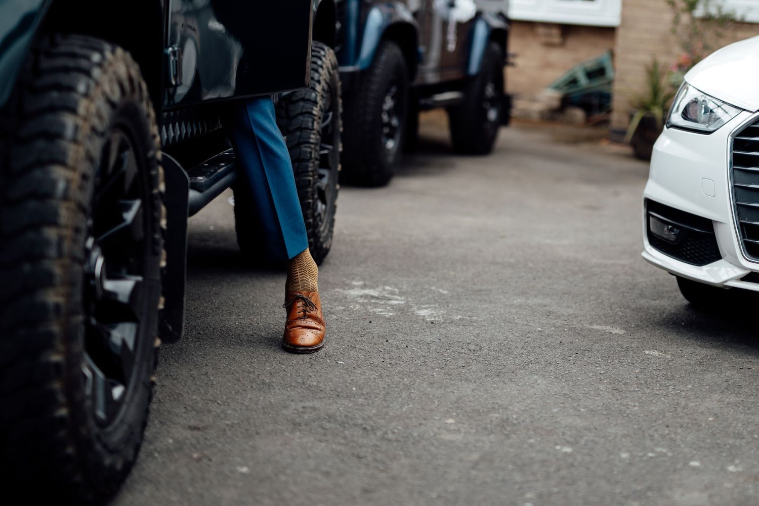 Brown leather boots stand between a motorcycle tire and car on concrete ground.
