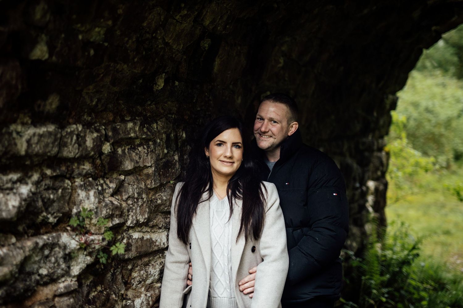 A couple shares an intimate moment under a stone archway in nature.