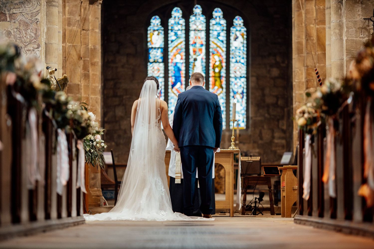 A wedding ceremony in a historic church with stained glass windows and floral decorations along the pews.