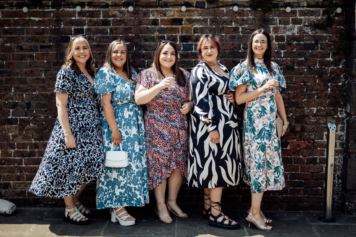 Group of wedding guests in floral dresses pose against a rustic brick wall.