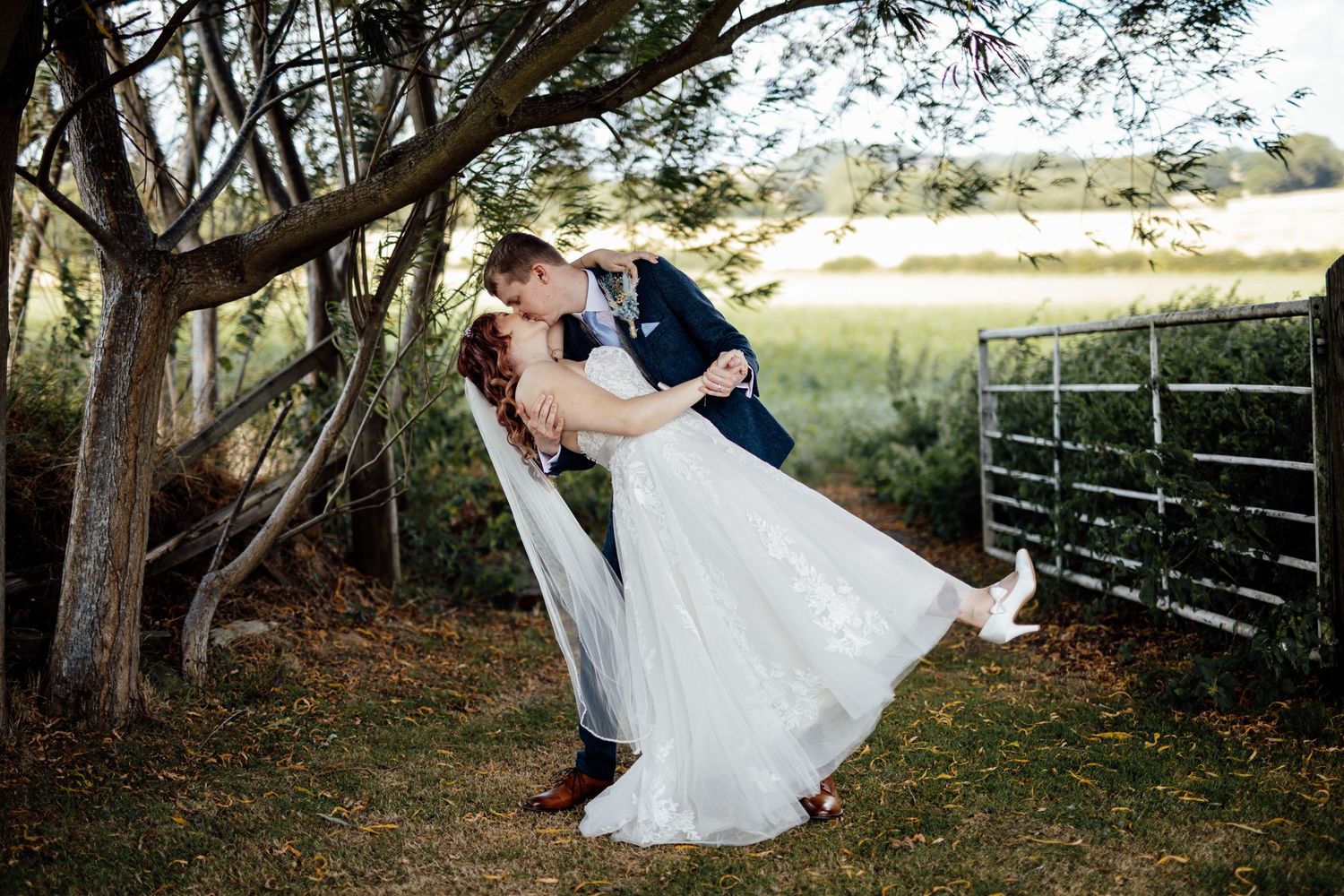 A romantic dip pose between newlyweds under trees near a wooden fence.