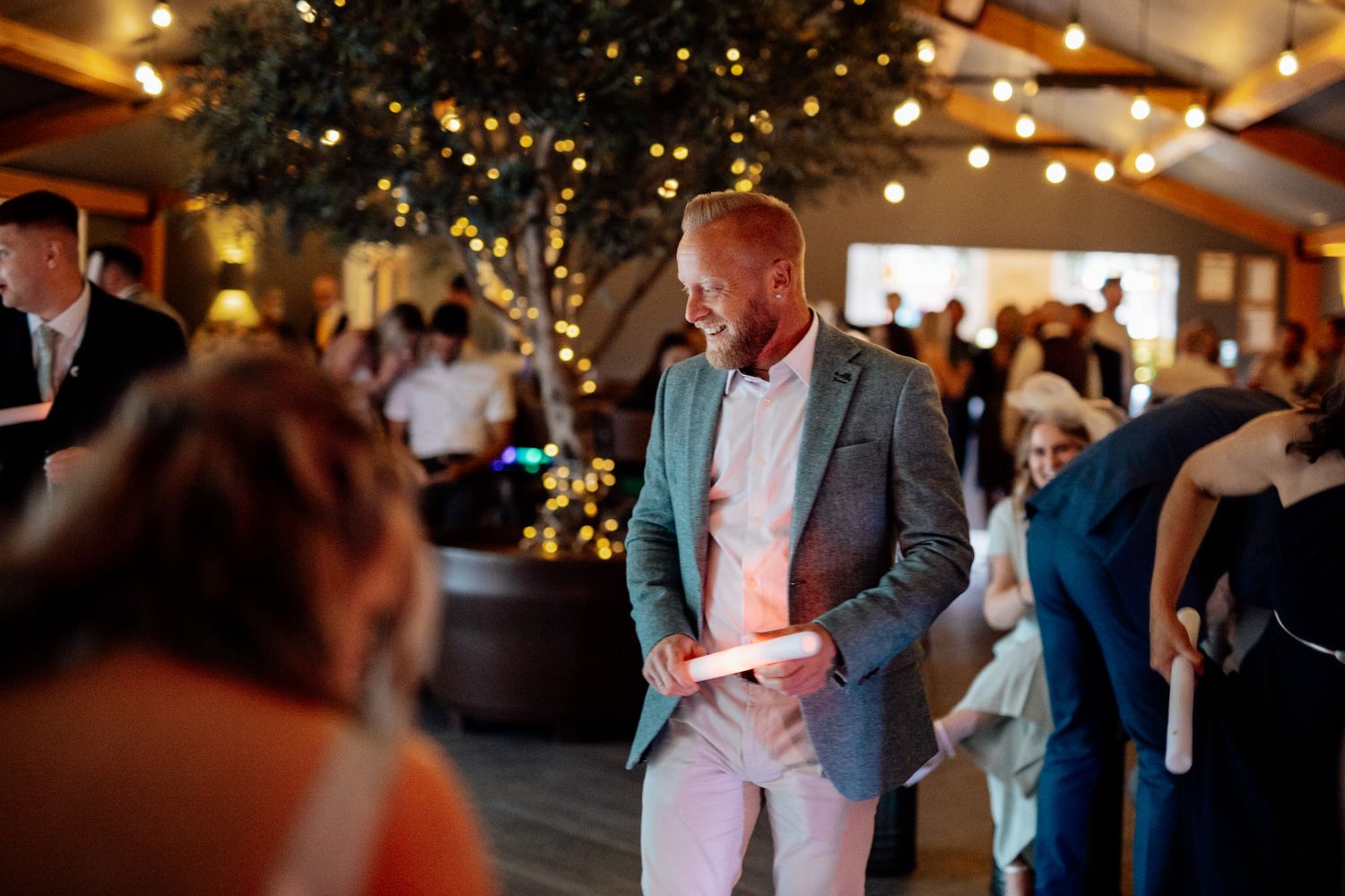 A guest dances at an indoor wedding reception venue decorated with string lights and greenery.