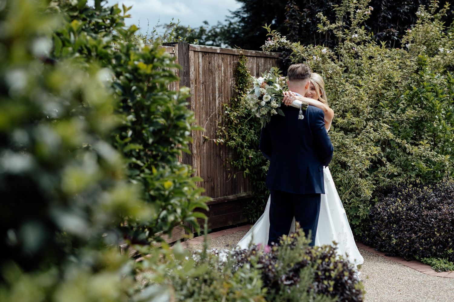 Wedding party poses in a lush garden setting with greenery and wooden fence backdrop.