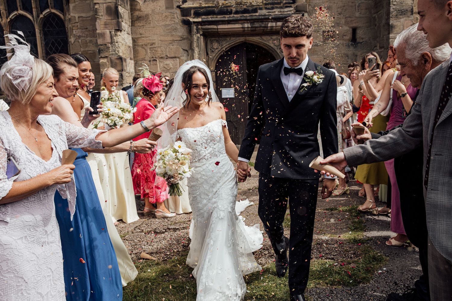 Wedding party celebrates with confetti toss outside historic stone church building.
