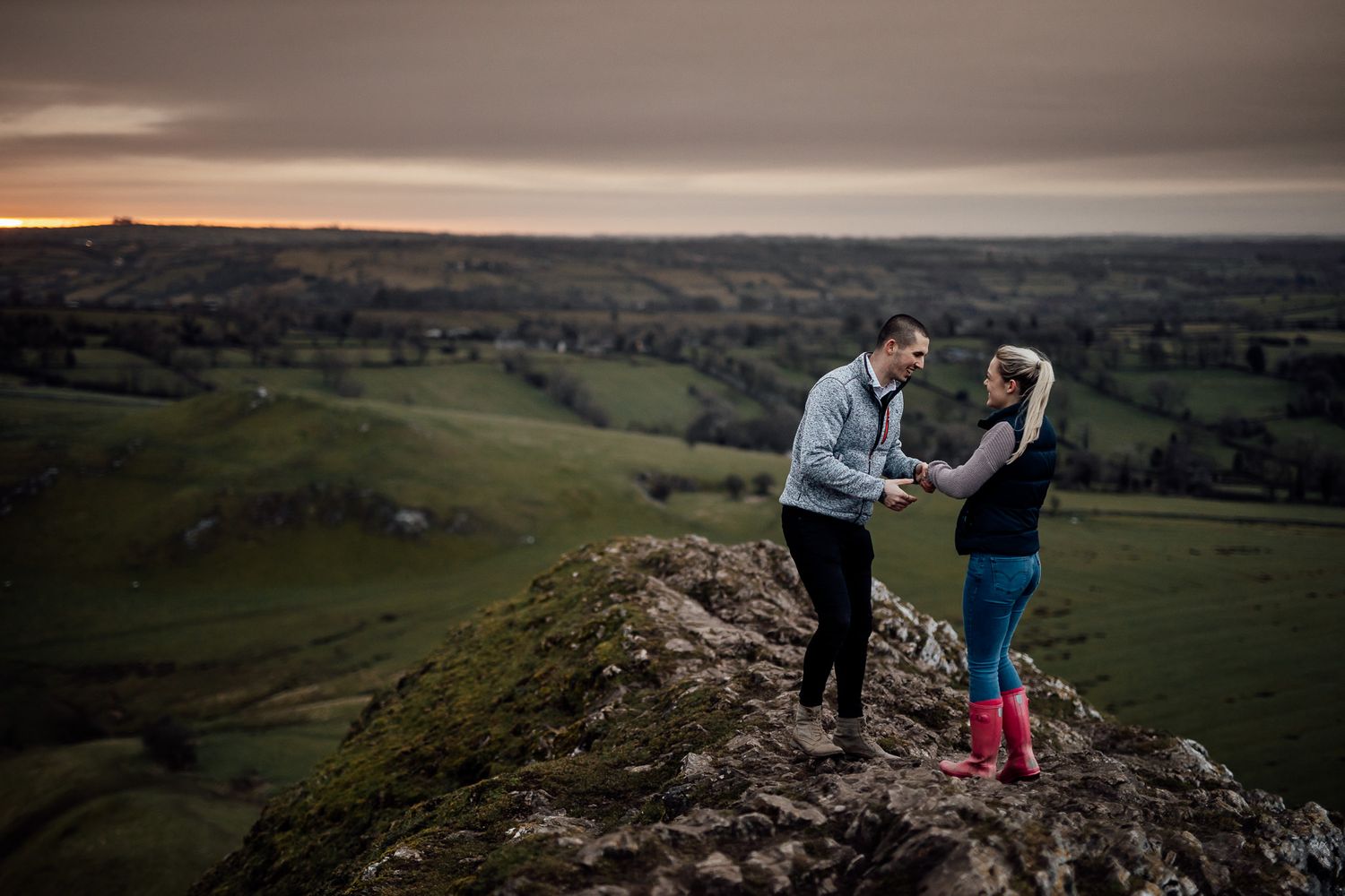Two people stand together on a rocky hilltop overlooking a vast valley landscape.