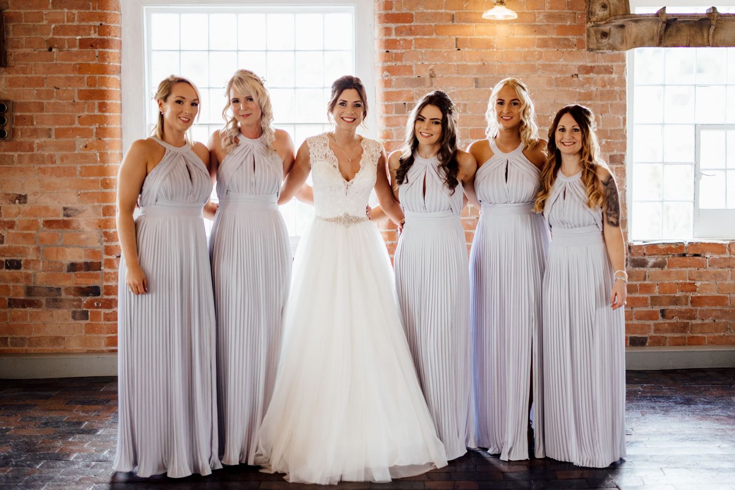 Wedding party poses in elegant gray bridesmaid dresses against exposed brick wall with natural window light.