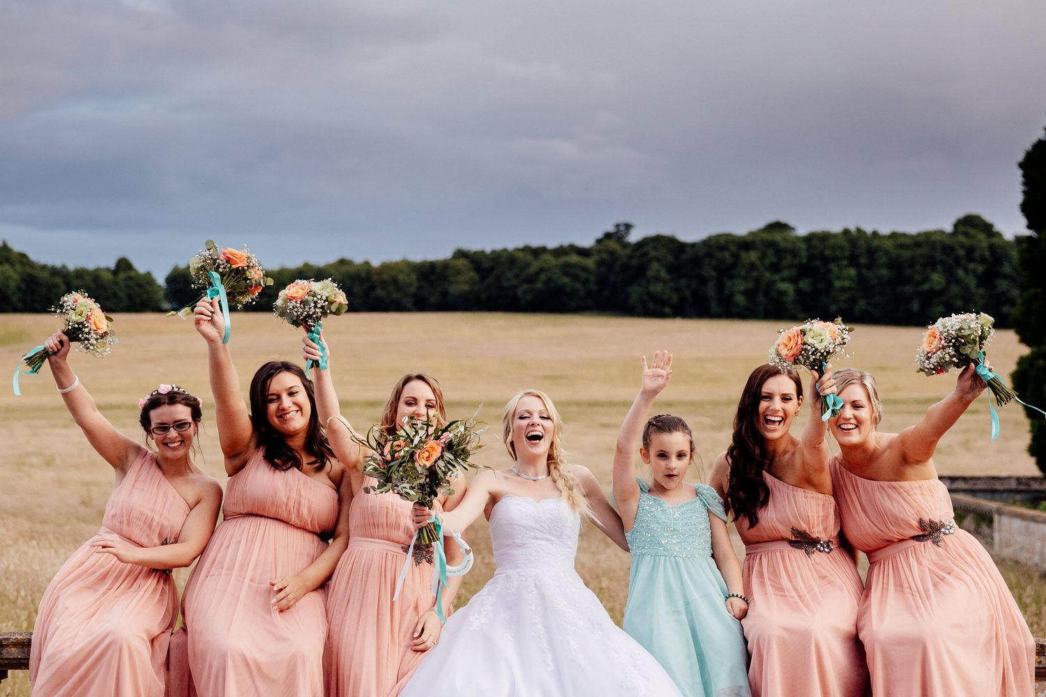 Bridal party in pink dresses poses joyfully on sunny lawn.