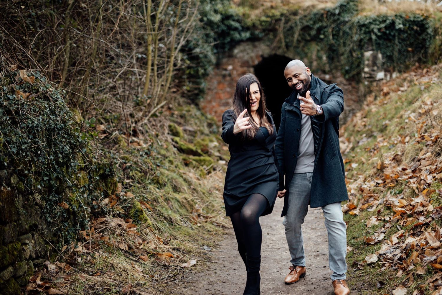 Two people stroll hand in hand along a rustic trail surrounded by fallen leaves and natural scenery.
