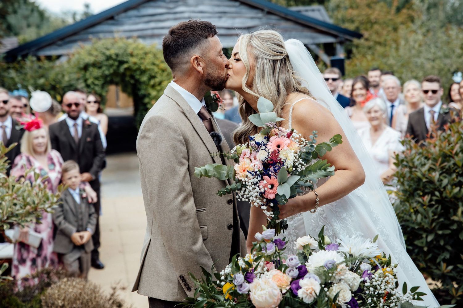Bride and groom share first kiss at outdoor wedding ceremony with guests celebrating behind them.