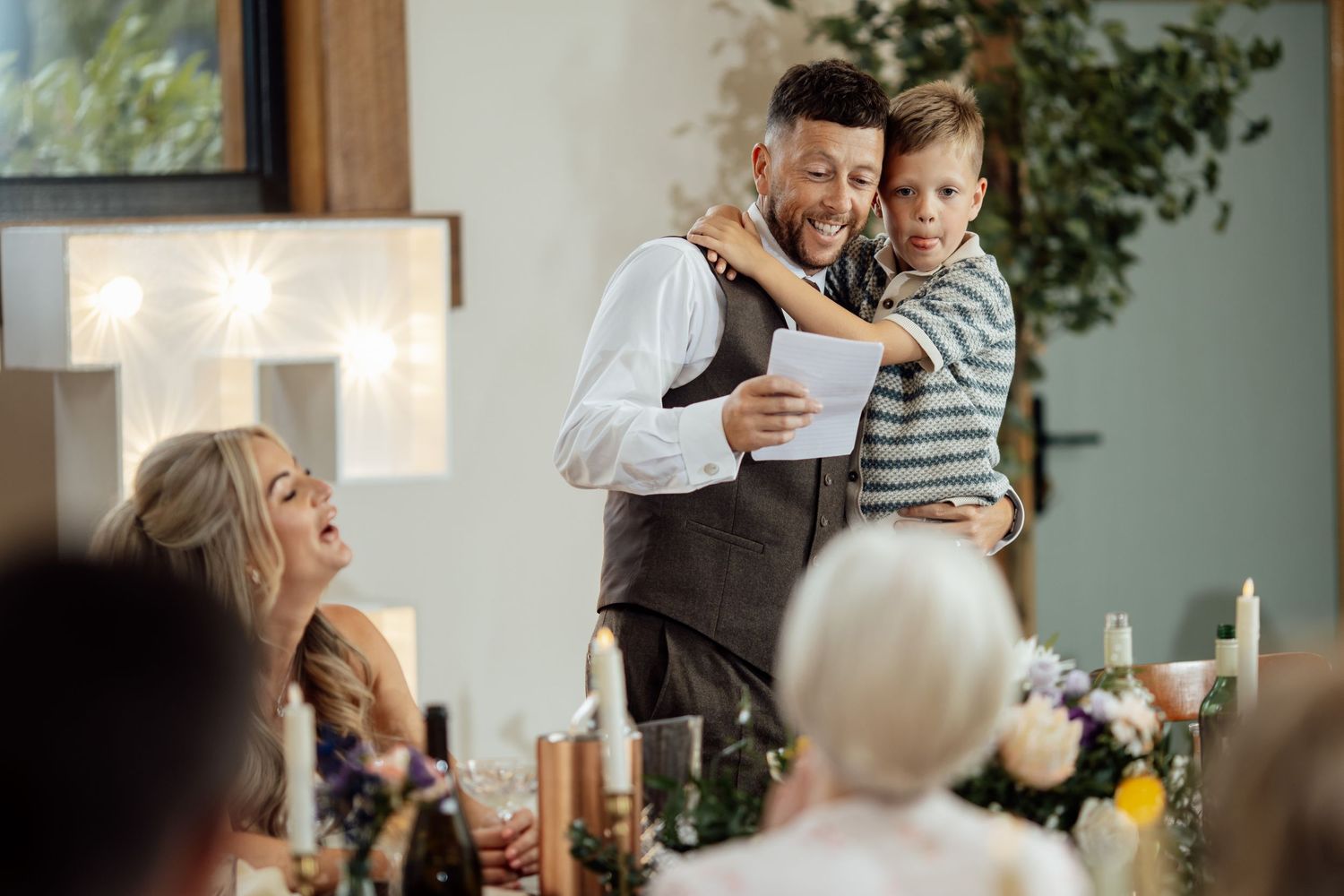 Smiling groom holding young boy while giving speech at wedding reception dinner with guests watching.