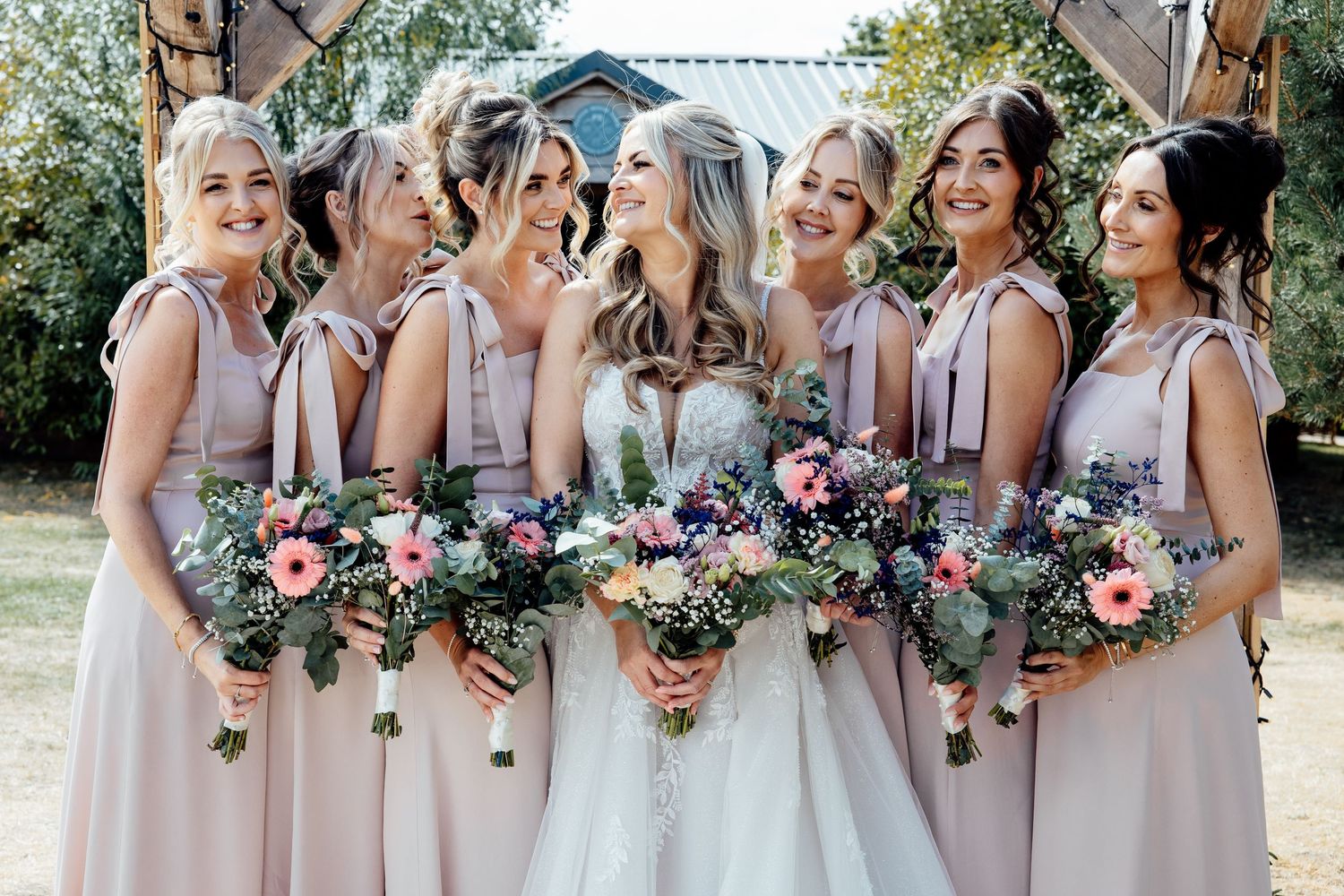 Bride poses with six bridesmaids in blush pink dresses holding colorful floral bouquets outdoors.