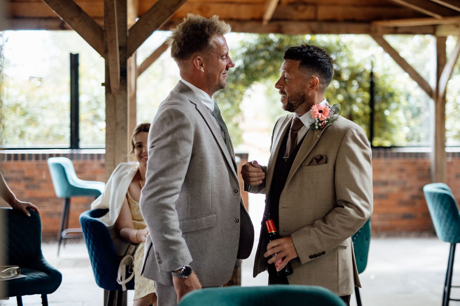 Groom in tan suit shaking hands with guest in modern wedding venue with teal chairs.