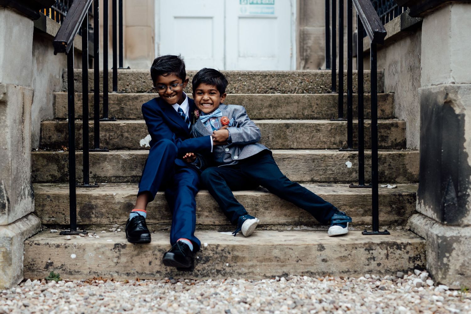Two young boys in blue suits sit playfully on stone steps sharing a lighthearted moment together.