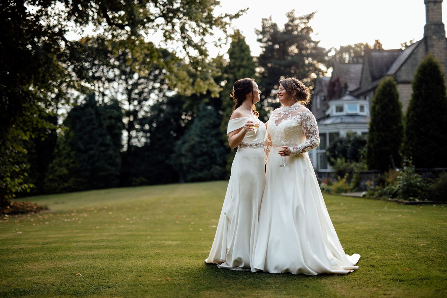 Two brides in white gowns share a joyful moment on a manicured lawn.