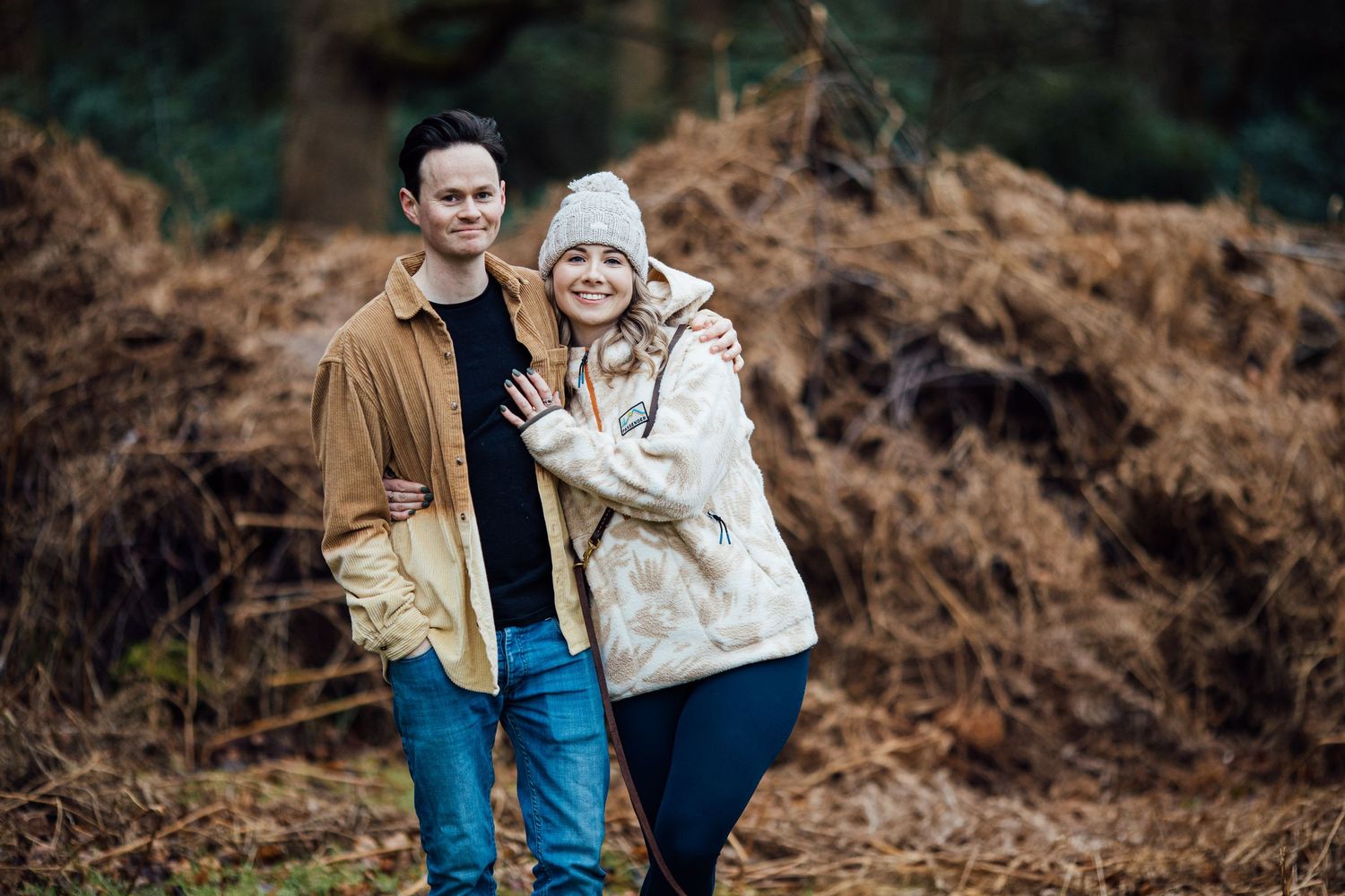 Couple in casual winter wear poses during engagement shoot in forest setting.