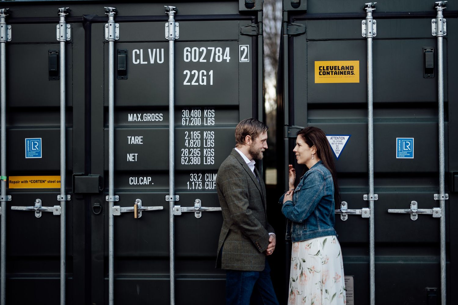 A couple shares an intimate moment against industrial shipping containers.