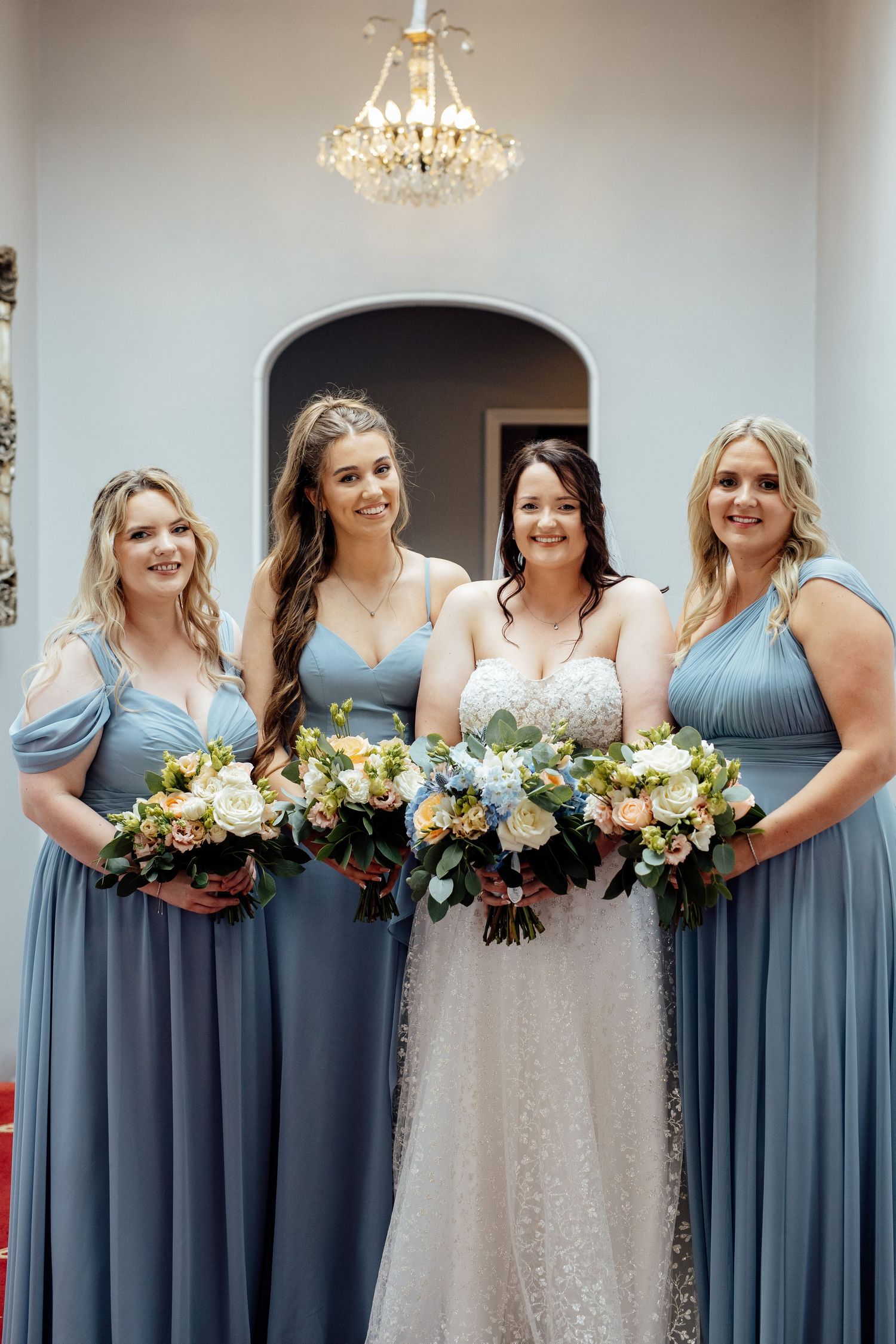 Bridal party in dusty blue dresses poses together under crystal chandeliers.