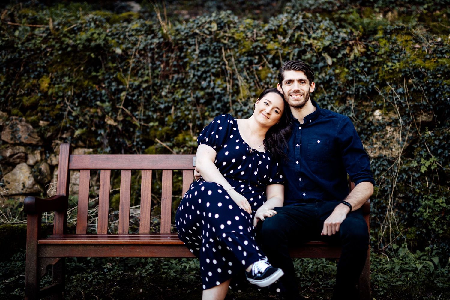 A couple relaxes on a wooden park bench surrounded by lush greenery.