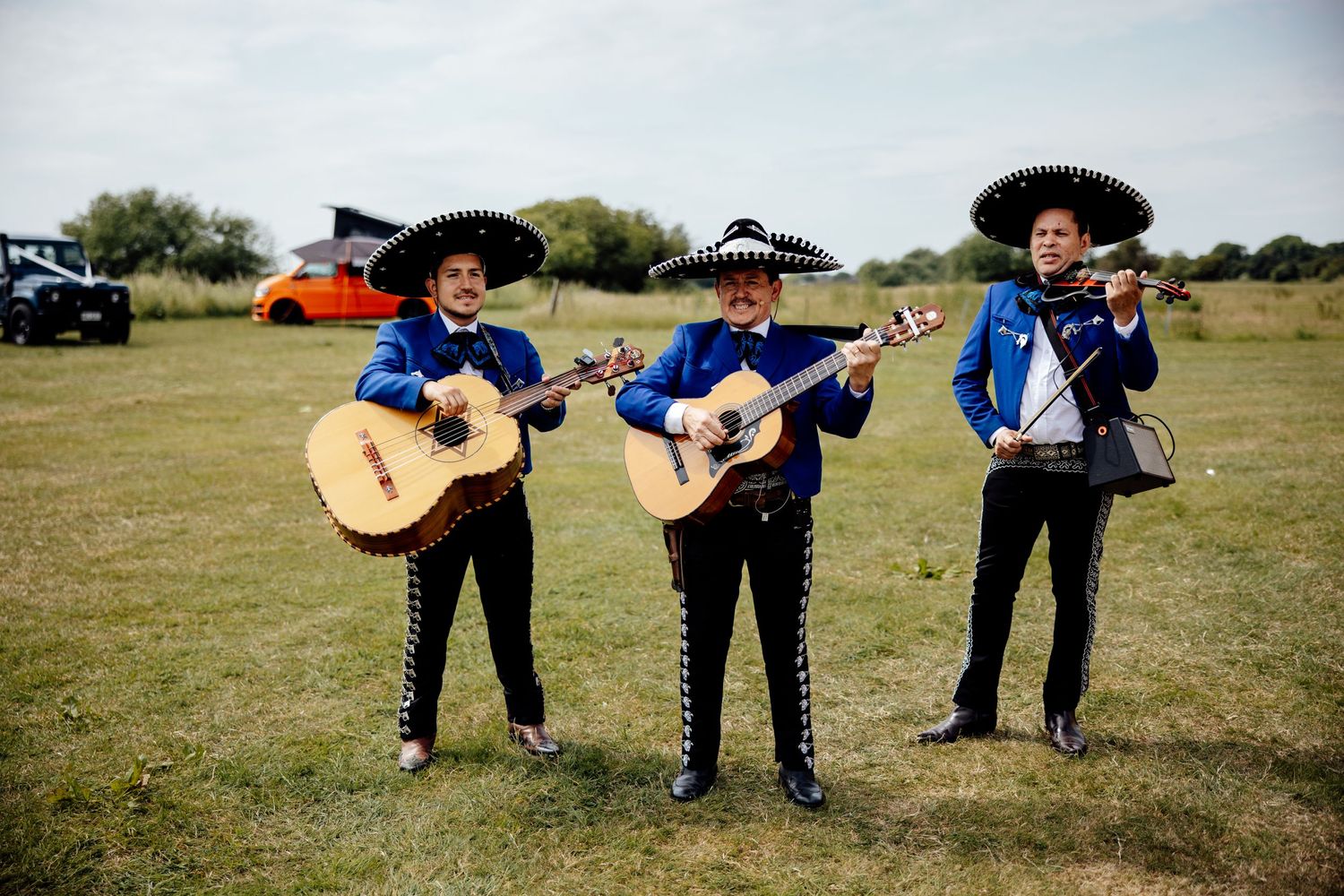 Three mariachi musicians in traditional sombreros and blue outfits perform with acoustic guitars on a grassy field.