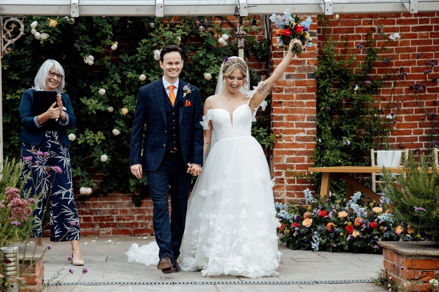 Wedding couple poses for photos in front of a rustic brick wall with string lights and greenery.
