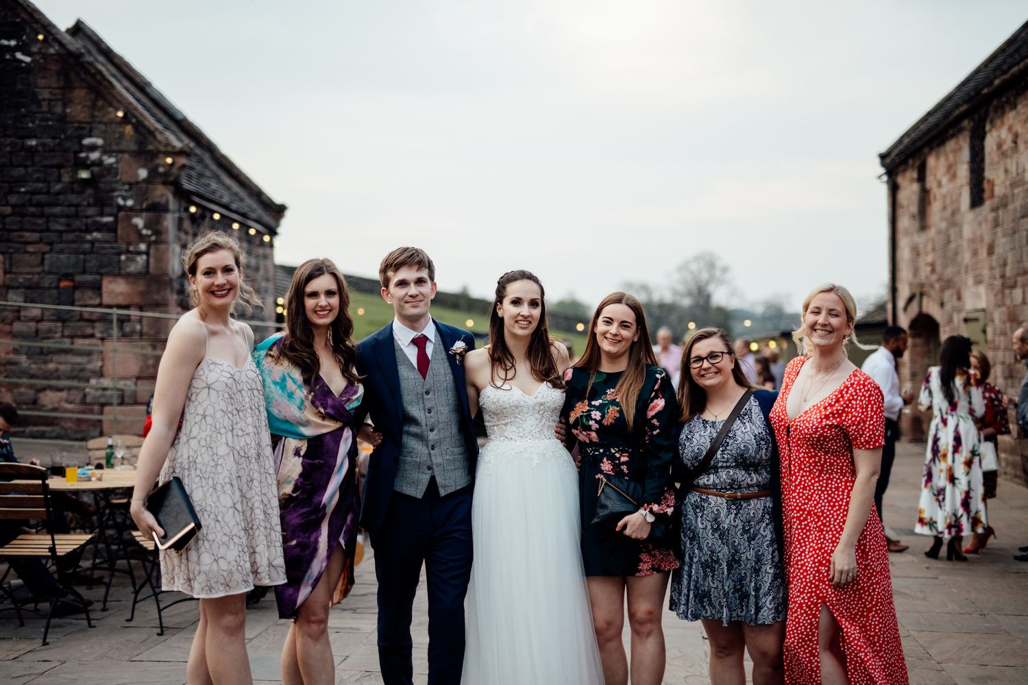 Wedding party and guests gather outside a rustic stone building for group photos.