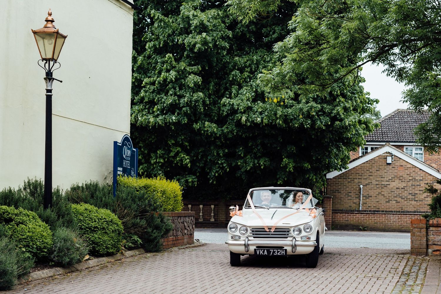 Vintage wedding car photographed along tree-lined street with historic buildings.