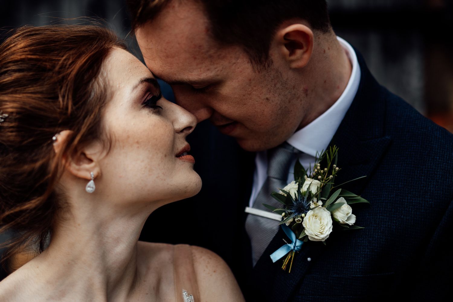 Intimate wedding couple moment captured in dramatic lighting with white floral boutonniere.