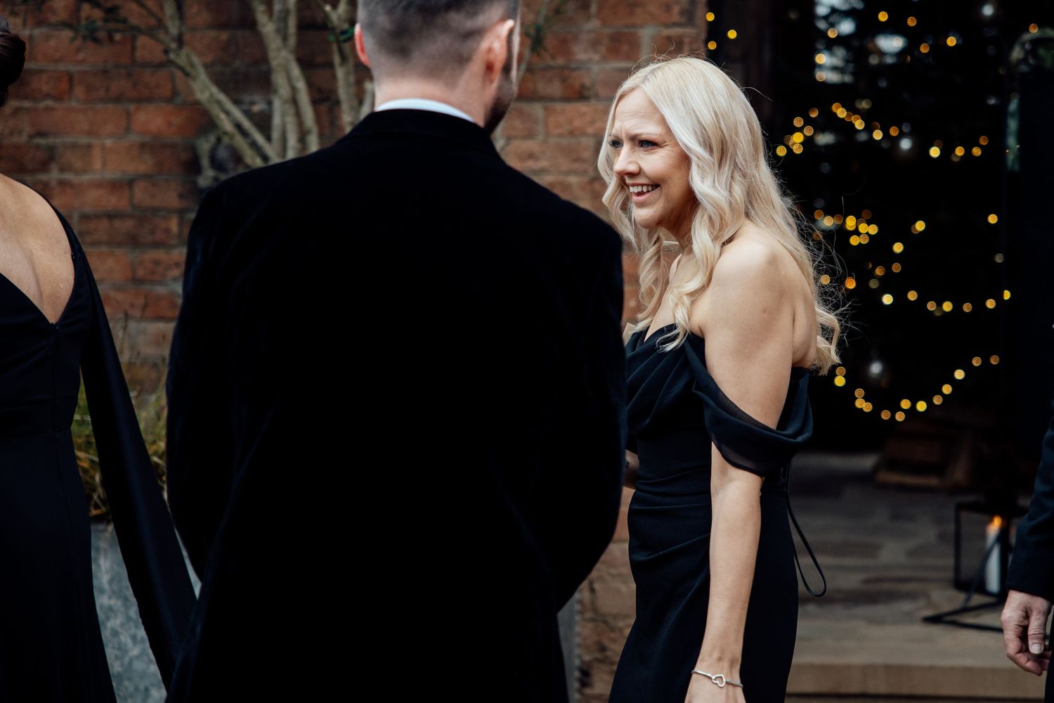 Wedding guest in a black formal dress engaged in conversation at an evening reception.