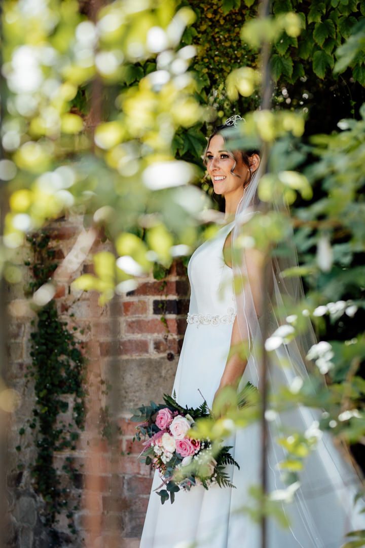 Bride in white dress holding pink bouquet poses against brick wall with soft natural lighting and foliage.