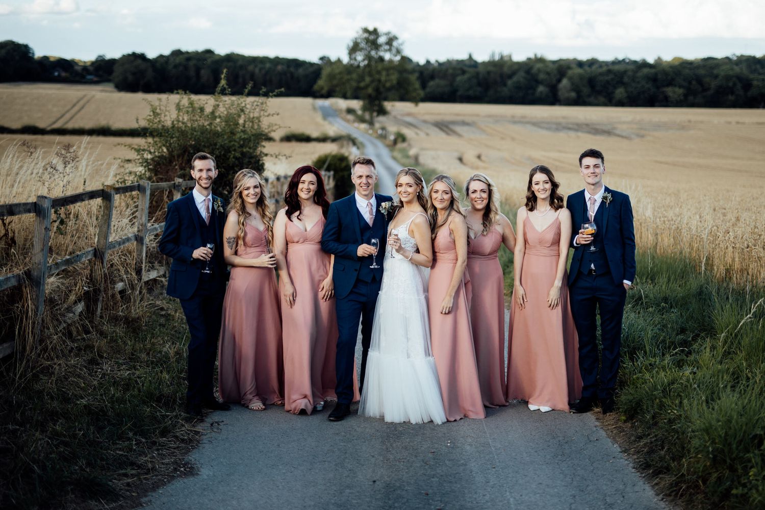 Wedding party stands together on rural path with wheat fields in background at dusk.