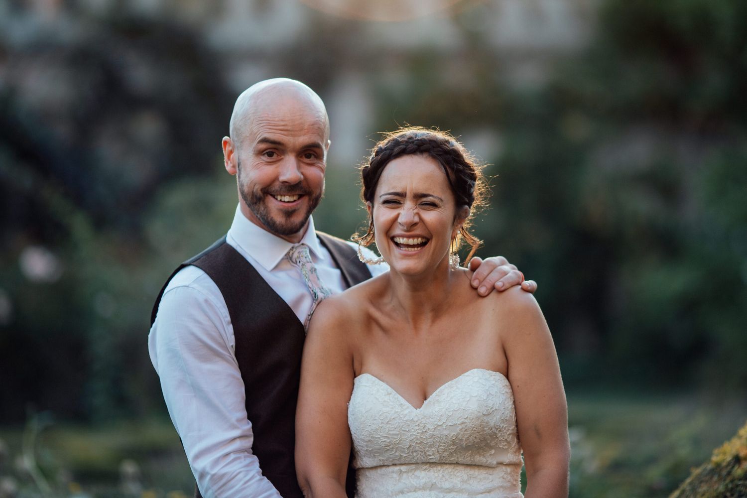 Joyful wedding couple embraces and laughs together in a romantic outdoor garden setting.