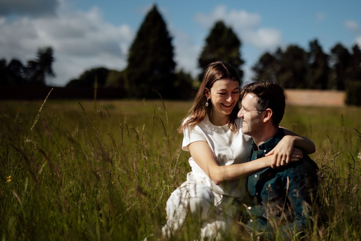 A couple embraces in a sunny meadow with trees and cloudy skies in the background.