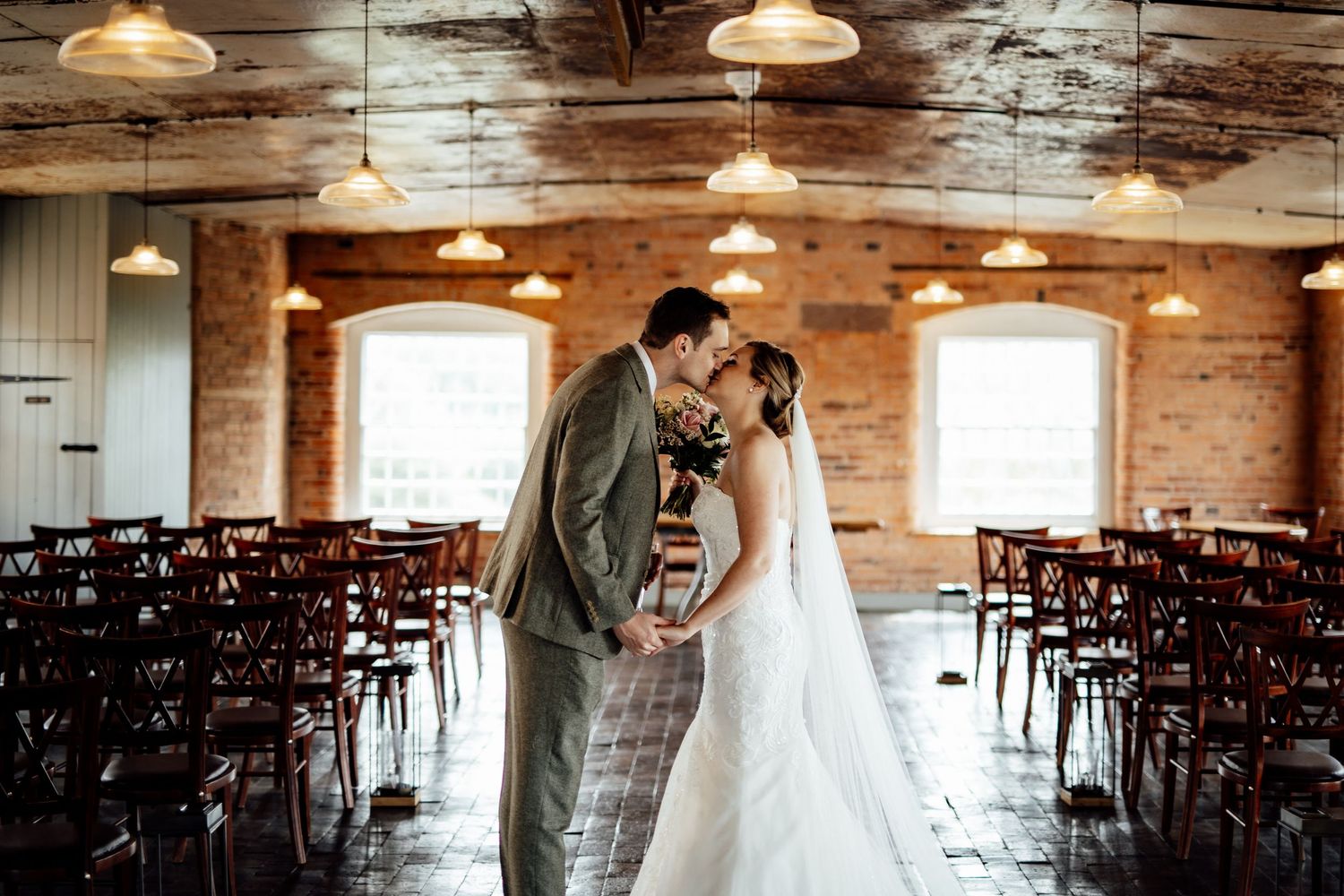 Rustic wedding venue interior with wooden beams and pendant lights arranged with rows of dark chairs.